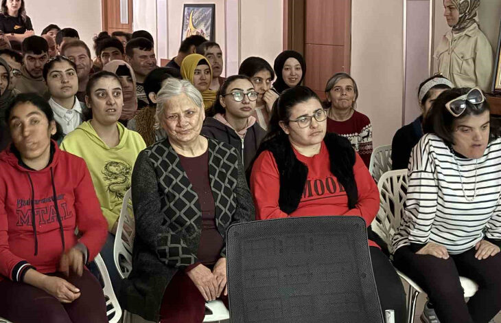 Görselde, bir grup insan bir salonda sandalyelere oturmuş, dikkatle bir etkinliği izliyor. Ön sırada yaşlı bir kadın, kırmızı kazaklı bir kadın ve çizgili bluz giyen bir başka kadın bulunuyor. Arkalarında ise farklı yaş gruplarından birçok kişi yer alıyor. Çoğu kişi ciddi ve odaklanmış bir ifadeyle dinliyor. Arka planda duvara asılmış tablolar ve ahşap kapılar göze çarpıyor.