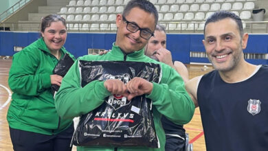 Fotoğrafta, bir spor salonunda çekilmiş mutlu bir an görülüyor. Ön planda gözlüklü, Down sendromlu bir genç yeşil spor kıyafetiyle elinde Beşiktaş logosu bulunan bir hediye paketi tutuyor ve gülümsüyor. Yanında siyah atlet giymiş bir adam da gülerek poz veriyor. Arkada ise başka bir genç kadın ve tekerlekli sandalyedeki bir sporcu bulunuyor. Tribünler boş, ortam spor salonunu andırıyor.