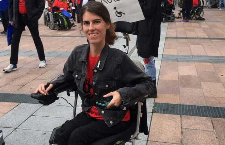 Görselde, tekerlekli sandalyede oturan bir kadın açık havada bir etkinliğe katılmaktadır. Kırmızı bir tişört ve siyah bir kot ceket giymiştir. Gülümseyerek kameraya bakmaktadır ve elinde bir telefon tutmaktadır. Arka planda, benzer renklerde giyinmiş ve tekerlekli sandalyede olan başka bireyler de dahil olmak üzere birçok kişi bulunmaktadır. Kadının arkasında, üzerinde tekerlekli sandalye simgesi bulunan bir pankart dikkat çekmektedir.