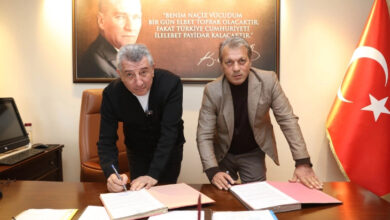 Fotoğrafta, bir ofis ortamında iki erkek bir masanın üzerinde bulunan belgeleri imzalıyor. Sol taraftaki kişi siyah bir ceket giyerken, sağdaki kişi açık kahverengi bir ceket içinde. Masanın üzerinde kırmızı ve beyaz dosyalar bulunuyor. Arka planda büyük bir Atatürk portresi ve ünlü bir sözü yer alıyor. Sağ tarafta ise Türk bayrağı bulunuyor.