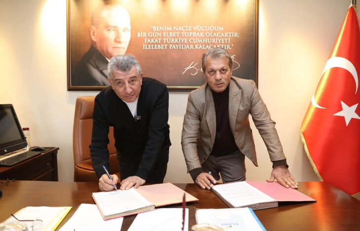 Fotoğrafta, bir ofis ortamında iki erkek bir masanın üzerinde bulunan belgeleri imzalıyor. Sol taraftaki kişi siyah bir ceket giyerken, sağdaki kişi açık kahverengi bir ceket içinde. Masanın üzerinde kırmızı ve beyaz dosyalar bulunuyor. Arka planda büyük bir Atatürk portresi ve ünlü bir sözü yer alıyor. Sağ tarafta ise Türk bayrağı bulunuyor.