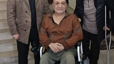 Görselde, tekerlekli sandalyede oturan bir adam, yanında iki kişiyle birlikte poz veriyor. Biri koltuk değneği kullanıyor, diğeri ise ayakta duruyor. Üçü de iç mekânda, bir merdivenin önünde duruyor. Gülümseyen yüz ifadeleri, samimi ve destekleyici bir ortamı yansıtıyor.