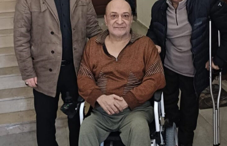 Görselde, tekerlekli sandalyede oturan bir adam, yanında iki kişiyle birlikte poz veriyor. Biri koltuk değneği kullanıyor, diğeri ise ayakta duruyor. Üçü de iç mekânda, bir merdivenin önünde duruyor. Gülümseyen yüz ifadeleri, samimi ve destekleyici bir ortamı yansıtıyor.
