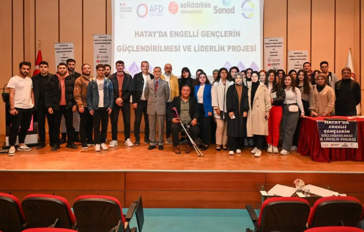 Görselde, bir konferans salonunda düzenlenen etkinlikte büyük bir grup insan sahnede toplu fotoğraf çektiriyor. Arkada büyük bir ekranda "Hatay’da Engelli Gençlerin Güçlendirilmesi ve Liderlik Projesi" yazısı ve çeşitli kuruluşların logoları yer alıyor. Katılımcılar arasında engelli bireyler ve destekçileri bulunuyor. Önde bir kişi tekerlekli sandalyede oturuyor ve elinde koltuk değneği var. Sol tarafta Türk bayrağı, sağda ise etkinlikle ilgili bilgiler içeren bir masa görülüyor.