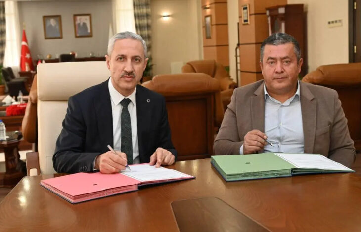 Görselde, resmi bir ofis ortamında iki erkek yan yana oturmuş ve bir protokol imzalamaktadır. Sol taraftaki kişi koyu renk takım elbise ve kravat giymiş, elinde kalemle belgeyi imzalarken kameraya bakmaktadır. Sağdaki kişi ise açık renk bir gömlek ve kahverengi bir ceket giymiş olup gözlüklerini elinde tutmaktadır. Masada kırmızı ve yeşil dosyalar bulunmaktadır.