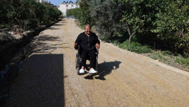 Görselde, tekerlekli sandalyede oturan bir kişi, toprak ve çakıl kaplı bir yolda ilerlemektedir. Kişi siyah kıyafetler giymiş ve beyaz terlikler giymektedir. Yolun etrafı yeşil ağaçlar ve bitkilerle çevrilidir. Arka planda bazı binalar görünmektedir. Hava güneşli ve açık görünüyor.