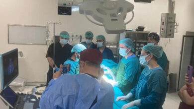 Görsel, bir ameliyathanede gerçekleştirilen tıbbi bir müdahaleyi gösteriyor. Ameliyat ekibi steril ameliyat önlükleri, eldivenler ve maskeler giymiş durumda. Parlak cerrahi ışıklar operasyon alanını aydınlatıyor. Çevrede birkaç kişi gözlem yapıyor, bazıları cep telefonlarıyla görüntü alıyor. Ekranlarda ultrason veya tıbbi görüntüler yer alıyor.