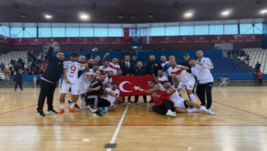 Görselde, kapalı bir spor salonunda bir grup sporcu ve teknik ekip, Türk bayrağıyla birlikte poz veriyor. Sporcular beyaz formalar giymiş ve sevinçlerini coşkuyla ifade ediyorlar. Arkada tribünlerde bazı izleyiciler yer alıyor. Takım, galibiyetin mutluluğunu paylaşırken birlik ve beraberlik duygusu ön plana çıkıyor.