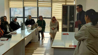 Görselde, bir sınıf ortamında eğitim verilen bir grup kadın görülüyor. Kadınların çoğu başörtülü ve kışlık kıyafetler giymiş, masaların etrafında oturuyorlar. Karşılarında bir erkek eğitmen, elinde kırmızı bir kitap tutarak konuşma yapıyor. Ön planda ise sırtı kameraya dönük bir kadın var. Sınıfta büyük pencereler bulunuyor ve doğal ışık içeri süzülüyor.