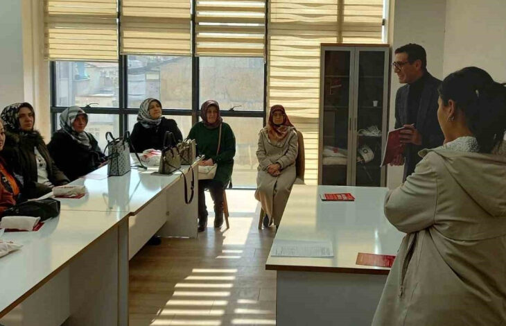 Görselde, bir sınıf ortamında eğitim verilen bir grup kadın görülüyor. Kadınların çoğu başörtülü ve kışlık kıyafetler giymiş, masaların etrafında oturuyorlar. Karşılarında bir erkek eğitmen, elinde kırmızı bir kitap tutarak konuşma yapıyor. Ön planda ise sırtı kameraya dönük bir kadın var. Sınıfta büyük pencereler bulunuyor ve doğal ışık içeri süzülüyor.