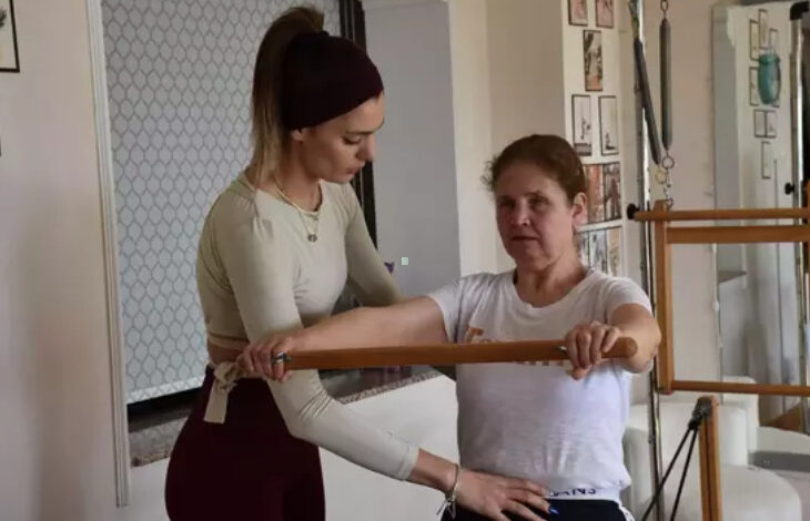 Görselde, bir spor salonunda veya pilates stüdyosunda iki kadın pilates egzersizi yapıyor. Beyaz tişörtlü, orta yaşlı bir kadın, ahşap bir çubuk kullanarak egzersiz yaparken yanında genç bir eğitmen ona yardımcı oluyor. Eğitmen, açık renk uzun kollu bir üst ve bordo bir tayt giymiş, saçları ise bantla toplanmış. Eğitmen, öğrencisinin doğru duruşu korumasına yardımcı olmak için ona fiziksel rehberlik sağlıyor. Arka planda ise çeşitli pilates ekipmanları ve duvarda asılı çerçeveler bulunuyor.