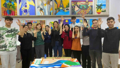 Görselde, bir grup genç ve öğretmenleri, renkli tabloların sergilendiği bir sanat atölyesinde poz veriyor. Grup, ellerini kaldırarak işaret diliyle bir mesaj veriyor olabilir. Masada yarım kalmış, canlı renklere sahip bir resim bulunuyor. Arka planda, doğa manzaraları ve soyut figürlerin yer aldığı çeşitli tablolar duvarda asılı.