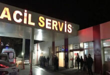 Fotoğrafta, gece vakti bir hastanenin acil servis girişi görülüyor. Büyük harflerle yazılmış "ACİL SERVİS" tabelası parlak ışıklarla aydınlatılmış. Kapının önünde birkaç kişi bekliyor, bazıları ayakta duruyor. Bir ambulans, acil servis girişinin yakınında park etmiş, mavi tepe lambası yanıyor.
