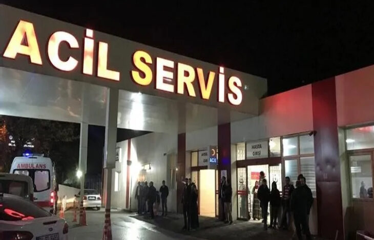 Fotoğrafta, gece vakti bir hastanenin acil servis girişi görülüyor. Büyük harflerle yazılmış "ACİL SERVİS" tabelası parlak ışıklarla aydınlatılmış. Kapının önünde birkaç kişi bekliyor, bazıları ayakta duruyor. Bir ambulans, acil servis girişinin yakınında park etmiş, mavi tepe lambası yanıyor.