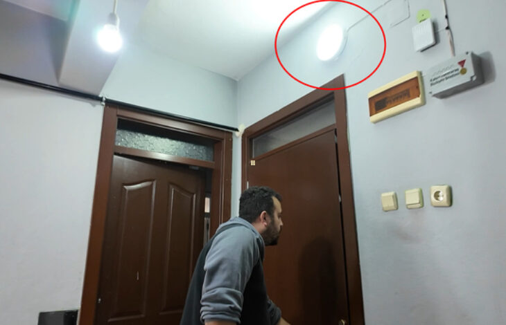 Görselde, kapı girişinde bulunan bir adam, yukarıya doğru bakıyor. Üst köşede, parlak bir ışık yayan bir lamba veya ışıklı zil dikkat çekiyor (kırmızı daire ile işaretlenmiş). Duvarlarda elektrik bağlantıları ve bazı anahtarlar bulunuyor. Ortam kapalı bir alan olup, kapılar kahverengi ahşap görünümlüdür.