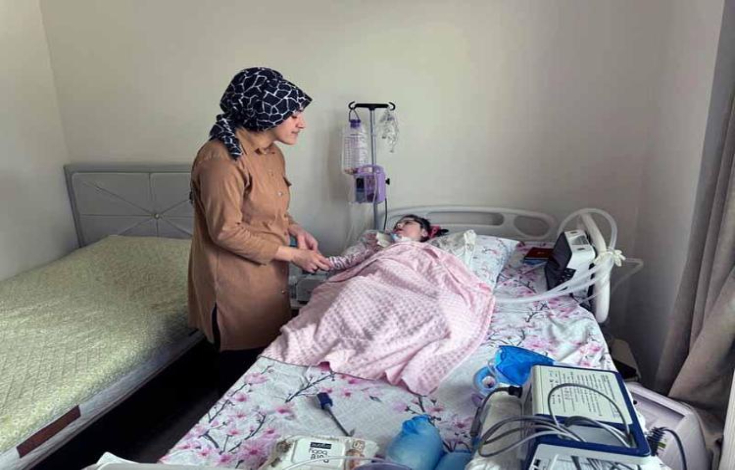 SMA'lı Elanur cihazlara bağlı yatağında uzanıyor ve annesi ayakta durarak elini tutuyor.