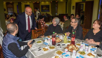 Fotoğrafta, bir iftar programında bir araya gelen insanlar görülüyor. Masalarda iftar yemekleri, ekmekler, içecekler ve yoğurt kapları bulunuyor. Takım elbiseli bir adam ayakta durarak masadaki kişilerle sohbet ediyor. Kadınlı erkekli bir grup insan, sıcak bir atmosfer içinde iftar yapmaya hazırlanıyor. Arka planda başka misafirler de yemeklerini yiyor ve sohbet ediyor.