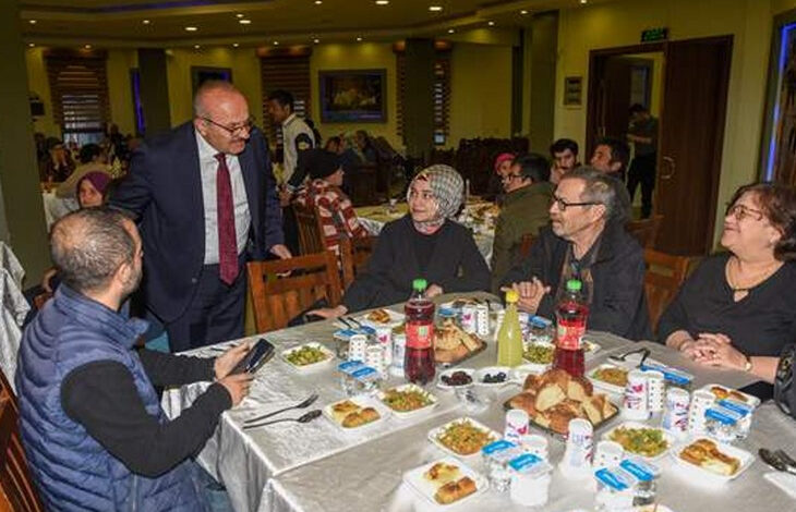 Fotoğrafta, bir iftar programında bir araya gelen insanlar görülüyor. Masalarda iftar yemekleri, ekmekler, içecekler ve yoğurt kapları bulunuyor. Takım elbiseli bir adam ayakta durarak masadaki kişilerle sohbet ediyor. Kadınlı erkekli bir grup insan, sıcak bir atmosfer içinde iftar yapmaya hazırlanıyor. Arka planda başka misafirler de yemeklerini yiyor ve sohbet ediyor.