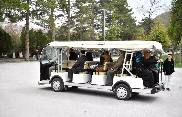 Görselde, üstü tente ile kaplı, açık yanları olan bir elektrikli servis aracı görülmektedir. Araçta çeşitli yaş grubundan insanlar oturmakta, bazıları başörtülü ve çoğunlukla koyu renkli kıyafetler giymektedir. Aracın arka tarafında bir kadın tutunarak ayakta duruyor. Şoför bölümü önde yer almakta. Arka planda ağaçlık bir alan, yürüyen insanlar ve mezar taşları seçilebiliyor, bu da buranın bir mezarlık olduğunu düşündürüyor.