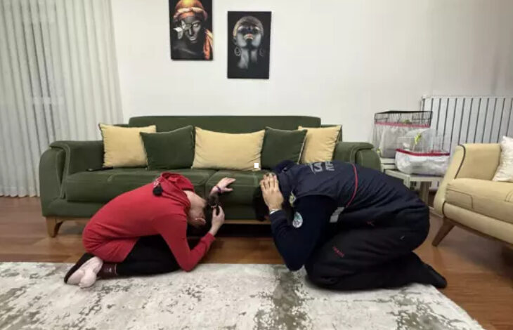 Görselde, bir ev ortamında iki kişi diz çökerek başlarını koruma pozisyonuna almış halde görülüyor. Kırmızı kıyafetli bir kadın ve itfaiye üniforması giymiş bir görevli, olası bir afet anında alınması gereken önlemleri uygulamalı olarak gösteriyor. Arkada yeşil bir koltuk, dekoratif yastıklar ve duvarda sanatsal tablolar bulunuyor. Bu görüntü, itfaiye ekiplerinin zihinsel engelli bir bireye afet eğitimi verdiği bir anı yansıtıyor.