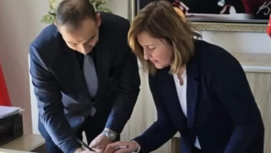 Görselde, bir ofis ortamında iki kişi bir belgeyi imzalamaktadır. Erkek olan kişi koyu renk takım elbise giymiş, kravat takmış ve belgeyi imzalarken eğilmiş durumdadır. Kadın ise lacivert bir ceket ve beyaz bir bluz giymiş olup, imza sürecine katılmaktadır. Masada pembe bir dosya bulunmaktadır. Arka planda çerçeveli bir tablo ve Türkiye bayrağı görünmektedir.