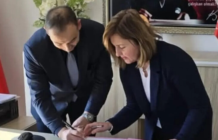 Görselde, bir ofis ortamında iki kişi bir belgeyi imzalamaktadır. Erkek olan kişi koyu renk takım elbise giymiş, kravat takmış ve belgeyi imzalarken eğilmiş durumdadır. Kadın ise lacivert bir ceket ve beyaz bir bluz giymiş olup, imza sürecine katılmaktadır. Masada pembe bir dosya bulunmaktadır. Arka planda çerçeveli bir tablo ve Türkiye bayrağı görünmektedir.