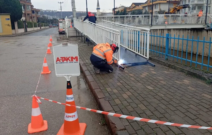 Görselde, Kocaeli Büyükşehir Belediyesi ekipleri tarafından Yahya Kaptan Mahallesi Akarca Köprüsü'nde yapılan engelli rampasının tamamlanma süreci görülüyor. İşçiler, güvenlik önlemleri alınmış bir alanda kaynak yaparak rampanın montajını gerçekleştiriyor. Çalışma alanı trafik konileri ve uyarı tabelaları ile çevrilmiş.