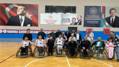 Görselde, bir spor salonunda toplanmış tekerlekli sandalye basketbol takımı ve destekçileri görülüyor. Sporcular, beyaz formalarla tekerlekli sandalyelerinde poz verirken, yanlarında antrenörler, yöneticiler ve bazı destekçiler yer alıyor. Arka planda Atatürk'ün büyük bir portresi, Cumhurbaşkanı Recep Tayyip Erdoğan’ın yer aldığı bir afiş ve “Türkiye Cumhuriyeti Gençlik ve Spor Bakanlığı” yazılı bir pano bulunuyor. Ayrıca “Kadına Şiddet İnsanlık Suçudur” yazılı bir pankart dikkat çekiyor.
