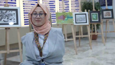 Görselde, başörtülü ve gözlük kullanan bir kadın, sanat eserlerinin sergilendiği bir alanda gülümseyerek kameraya poz veriyor. Açık renkli bir ceket giymiş olan kadın, boynunda desenli bir şal ve kimlik kartı taşıyor. Arkasında, çeşitli resimlerin sergilendiği ahşap şövaleler ve bir bilgilendirme panosu yer alıyor.