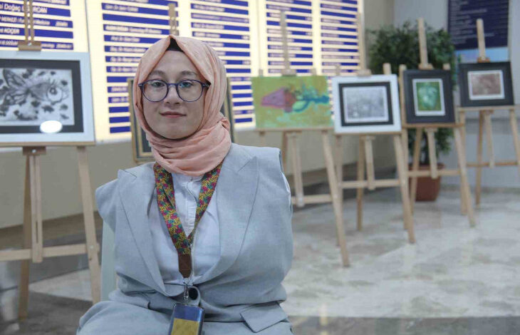 Görselde, başörtülü ve gözlük kullanan bir kadın, sanat eserlerinin sergilendiği bir alanda gülümseyerek kameraya poz veriyor. Açık renkli bir ceket giymiş olan kadın, boynunda desenli bir şal ve kimlik kartı taşıyor. Arkasında, çeşitli resimlerin sergilendiği ahşap şövaleler ve bir bilgilendirme panosu yer alıyor.