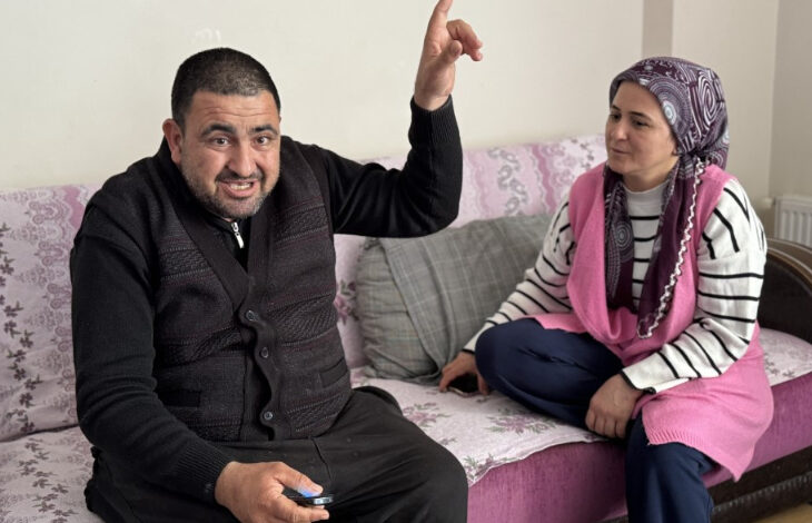 Görselde, bir kanepe üzerinde oturan iki kişi görülüyor. Soldaki adam koyu renk kıyafetler giymiş, sakallı ve elinde bir cep telefonu tutuyor. Sağ eliyle yukarıyı işaret ederken yüzünde konuşkan ve canlı bir ifade var. Sağdaki kadın ise başörtülü, pembe bir yelek giymiş ve onu dikkatlice dinlerken hafifçe gülümsüyor.