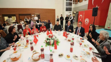 Görselde, büyük bir salonda yuvarlak masalarda düzenlenen bir iftar yemeği görülüyor. Masaların üzerinde Türk bayrakları, çiçek süslemeleri ve çeşitli yemekler bulunuyor. Katılımcılar dua ederken görüntülenmiş. Masada Muğla Valisi İdris Akbıyık ve eşi Sevim Akbıyık da yer alıyor. Arka planda büyük bir Türk bayrağı ve resmi pankartlar bulunuyor. Ayrıca, davetlilere hizmet eden garsonlar da dikkat çekiyor.