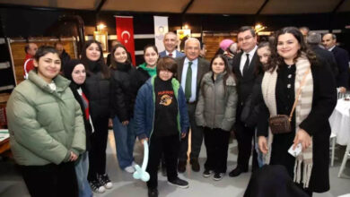 Görselde, bir grup insan kapalı bir mekânda bir araya gelmiş ve birlikte poz veriyor. Ön planda gençler ve kadınlar yer alırken, arka planda takım elbiseli birkaç kişi dikkat çekiyor. Katılımcılar arasında yaşça büyük bir adam da bulunuyor ve yanında resmi giyimli kişiler var. Arkada Türk bayrağı ve çeşitli afişler asılı.