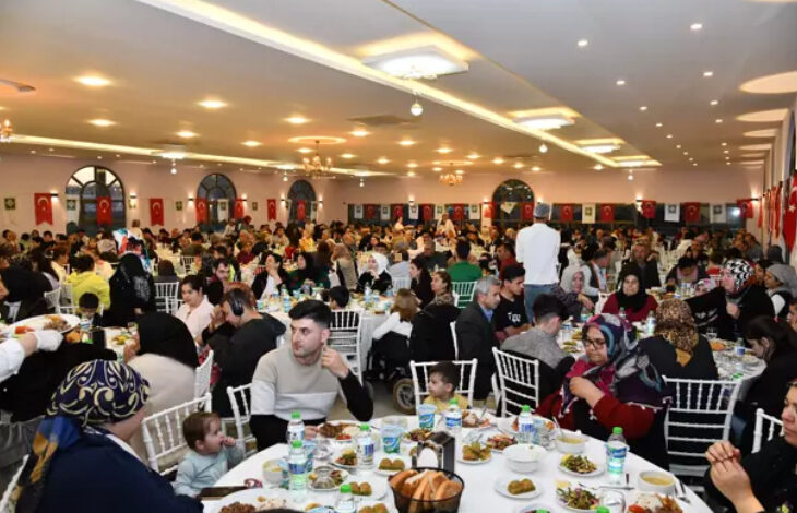 Görsel, büyük bir salonda düzenlenen kalabalık bir iftar yemeğini gösteriyor. Yuvarlak masalarda oturan çok sayıda insan iftarlarını açıyor. Masalarda çeşitli yemekler, ekmekler ve su şişeleri bulunuyor. Katılımcılar arasında kadınlar, erkekler ve çocuklar yer alıyor. Duvarlarda Türk bayrakları asılı ve ortam oldukça aydınlık.