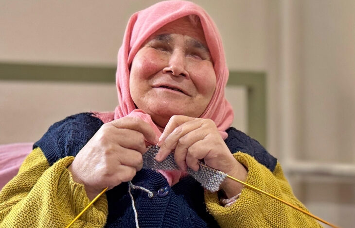 Görselde, pembe başörtüsü takan yaşlı bir kadın, elinde şiş ve iplerle örgü örüyor. Üzerinde sarı kollu kalın bir kazak ve lacivert bir yelek bulunuyor. Yüzünde hafif bir gülümseme var ve gözleri tamamen kapalı gibi görünüyor. Arka plan nötr tonlarda ve iç mekânda çekilmiş bir fotoğraf izlenimi veriyor.