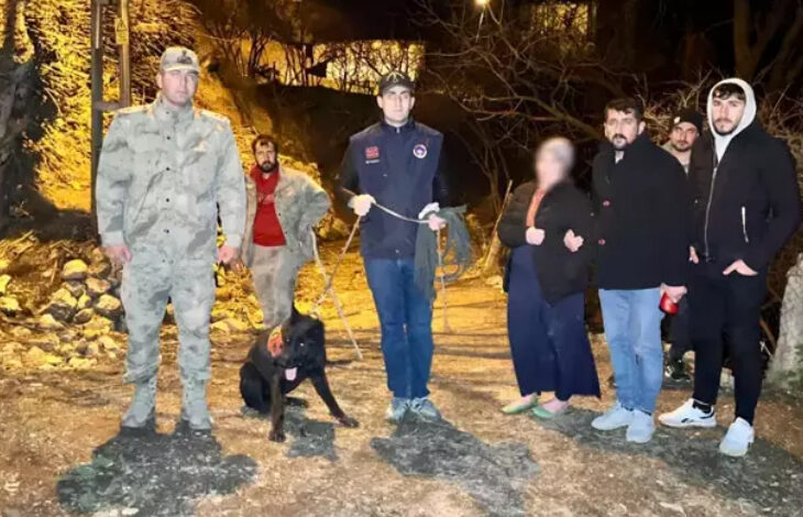 Görselde gece vakti açık alanda çekilmiş bir sahne yer alıyor. Ortada bir iz takip köpeği ve onu tutan bir görevli bulunuyor. Yanlarında askeri üniformalı bir kişi, sivil giyimli birkaç kişi ve bir kadın var. Kadının yüzü bulanıklaştırılmış. Çevre doğal ve ormanlık bir alan gibi görünüyor.