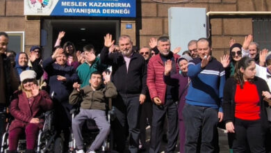 Görselde, bir grup insan, bir binanın önünde toplanmış ve kameraya el sallıyor. Binanın üzerinde "Zile Engellilere Meslek Kazandırma Dayanışma Derneği" yazılı bir tabela bulunuyor. Grupta tekerlekli sandalyede oturan bireyler de dahil olmak üzere farklı yaş gruplarından insanlar yer alıyor. Katılımcılar neşeli ve dayanışma içinde görünüyor.