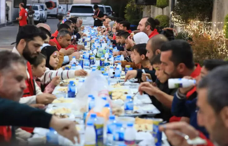 Görselde, uzun bir masada açık havada topluca yemek yiyen insanlar görülüyor. Masanın üzerinde su, ayran ve çeşitli yemeklerin bulunduğu tepsiler yer alıyor. Katılımcılar arasında çocuklar ve yetişkinler var, bazı kişiler Kızılay yeleği giymiş durumda. Arka planda ışık süslemeleri ve park edilmiş araçlar dikkat çekiyor.