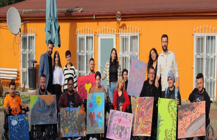 Görselde, açık hava ortamında bir grup insan birlikte poz vermektedir. Ön sırada tekerlekli sandalyede oturan bireyler ellerinde rengarenk resimler tutmaktadır. Arkada ise ayakta duran birkaç kişi bulunmaktadır. Grup, mutlu ve enerjik bir şekilde kameraya bakmaktadır. Arkada turuncu renkli bir bina, beyaz pencereler ve bir uydu anteni görülmektedir.