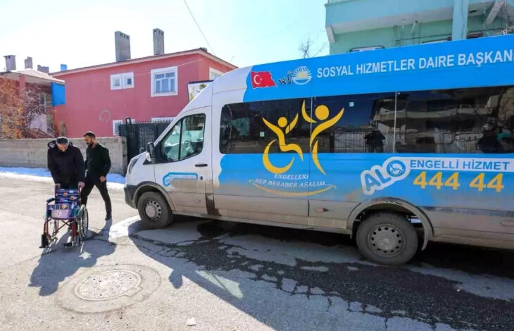 Görselde, Van Büyükşehir Belediyesi'ne ait bir engelli hizmet aracı görülüyor. Aracın üzerinde "Sosyal Hizmetler Daire Başkanlığı" ve "Engelleri Hep Beraber Aşalım" yazıları yer alıyor. Ayrıca, iletişim hattı olarak "ALO Engelli Hizmet 444 44" numarası bulunuyor. Aracın yanında, bir kişi tekerlekli sandalye ile yürürken yanında bir başka kişi ona eşlik ediyor. Arka planda renkli evler ve açık bir hava görülüyor.