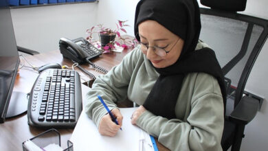 Görselde, bir kadın ofis ortamında masa başında çalışmaktadır. Siyah başörtüsü ve yeşil bir sweatshirt giymiştir. Gözlük takmakta ve elinde bir kalemle kağıt üzerine yazı yazmaktadır. Masasında bir bilgisayar klavyesi, telefon, bazı belgeler ve küçük bir saksı bitkisi bulunmaktadır. Arka planda mavi klasörlerin sıralandığı bir raf dikkat çekmektedir.