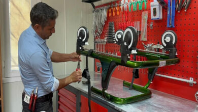 Bu görselde, bir tamir atölyesinde çalışan bir adam görülüyor. Adam, bir masa üzerinde ters çevrilmiş bir tekerlekli sandalye şasisine benzer bir yapıyı tamir ediyor. Üzerinde çeşitli tamir aletleri taşıyan bir pantolon giymiş. Arka planda kırmızı bir alet panosu bulunuyor ve üzerinde birçok pense, anahtar ve tornavida gibi tamir aletleri asılı.