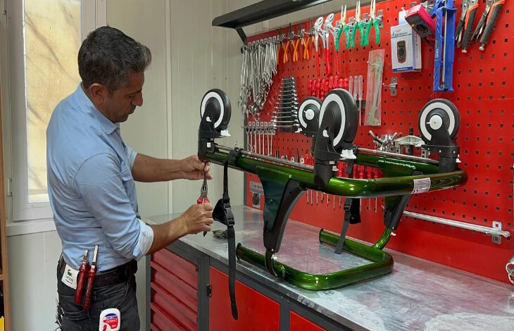 Bu görselde, bir tamir atölyesinde çalışan bir adam görülüyor. Adam, bir masa üzerinde ters çevrilmiş bir tekerlekli sandalye şasisine benzer bir yapıyı tamir ediyor. Üzerinde çeşitli tamir aletleri taşıyan bir pantolon giymiş. Arka planda kırmızı bir alet panosu bulunuyor ve üzerinde birçok pense, anahtar ve tornavida gibi tamir aletleri asılı.