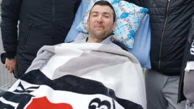 Görselde, bir sedyede yatan bir adam görülmektedir. Adam, üzerinde Beşiktaş Jimnastik Kulübü'nün logosunu taşıyan bir battaniyeye sarılmıştır. Başının altında mavi ve beyaz çiçek desenli bir yastık bulunmaktadır. Adamın yüzünde hafif bir gülümseme ifadesi vardır ve gözleri açık bir şekilde yukarıya bakmaktadır.