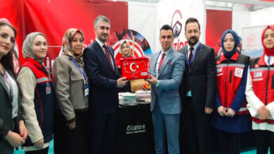 Bu görselde, bir fuar veya etkinlik alanında çekilmiş bir fotoğraf yer alıyor. Ortada iki erkek, Türkiye haritası şeklinde hazırlanmış ve Türk bayrağı motifli bir plaketi birlikte tutuyor. Çevrelerinde çeşitli kurum kıyafetleri giymiş kadınlar ve takım elbise giymiş erkekler bulunuyor. Arkada "Güzide Bakım Merkezleri" ve "Aile ve Sosyal Hizmetler Bakanlığı" logoları içeren afişler görülüyor.