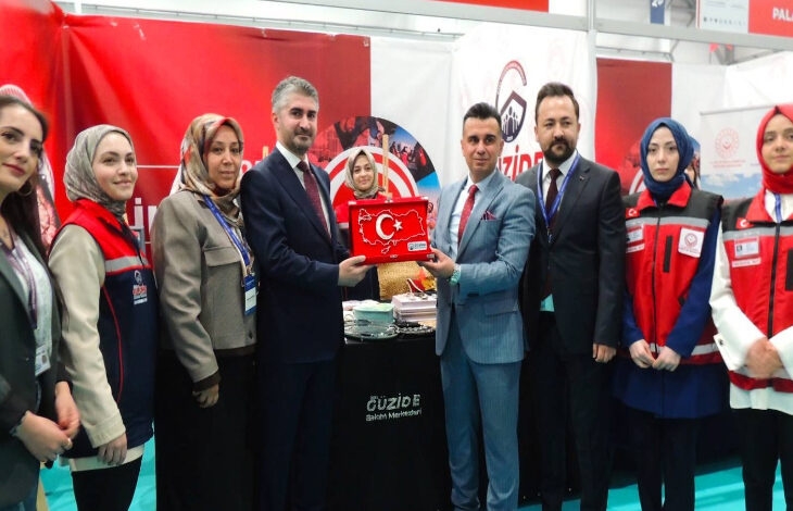 Bu görselde, bir fuar veya etkinlik alanında çekilmiş bir fotoğraf yer alıyor. Ortada iki erkek, Türkiye haritası şeklinde hazırlanmış ve Türk bayrağı motifli bir plaketi birlikte tutuyor. Çevrelerinde çeşitli kurum kıyafetleri giymiş kadınlar ve takım elbise giymiş erkekler bulunuyor. Arkada "Güzide Bakım Merkezleri" ve "Aile ve Sosyal Hizmetler Bakanlığı" logoları içeren afişler görülüyor.
