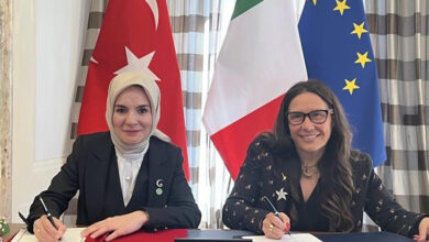 Bu görselde, iki kadın temsilci resmi bir belge imzalarken görülüyor. Sol tarafta yer alan kadın başörtülü ve koyu renkli resmi bir kıyafet giymiş. Sağ tarafta yer alan kadın ise gözlüklü ve yıldız desenli bir ceket giymekte. Arka planda Türkiye, İtalya ve Avrupa Birliği bayrakları yer alıyor. Bu detaylar, uluslararası bir iş birliğini veya protokol anlaşmasını temsil etmektedir.