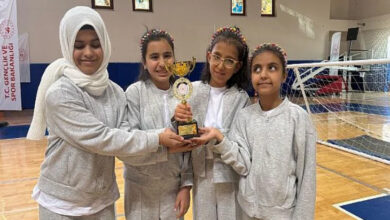 Bu görselde, bir spor salonunda gri eşofman takımları giymiş dört kız çocuğu yer almakta. Hepsi birlikte bir kupa tutuyorlar ve mutlular. Kızlardan biri başörtüsü takıyor, diğer üçü ise saçlarına renkli tokalar takmış. Arka planda "T.C. Gençlik ve Spor Bakanlığı" yazılı bir afiş ve fileli bir spor alanı görülüyor.