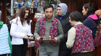 Bu fotoğrafta, geleneksel desenli yelek giymiş bir genç erkek elinde çay tepsisiyle servis yaparken görülüyor. Etrafında farklı yaşlardan kadınlar bulunmakta; bazıları onunla sohbet ediyor, bazıları ise çevreyi inceliyor. Arka planda çeşitli el işi süs eşyalarının ve nazar boncuklarının sergilendiği bir dükkân yer almakta. Mekân, kültürel ve turistik bir atmosfere sahip.