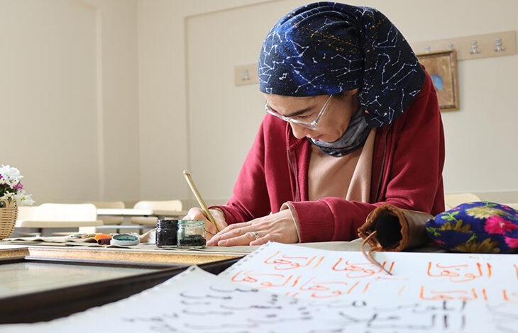 Görselde, başörtülü bir kadın hat sanatıyla uğraşırken görülüyor. Kırmızı bir hırka giymiş olan kadın, bir sınıf ortamında masaya oturmuş ve elinde bir hat kalemiyle dikkatlice yazı yazıyor. Masanın üzerinde mürekkep, kalem ve Arap harfleriyle yazılmış kâğıtlar bulunuyor. Arka planda ise boş sıralar ve duvarda asılı askılıklar mevcut. Kadının sanata olan ilgisi ve çalışmasına olan yoğun dikkati hissediliyor.